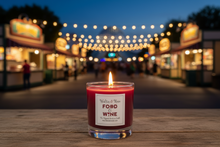 Load image into Gallery viewer, A lit red candle in a clear glass jar sits on a wooden surface with a blurred night market street in the background, featuring warm string lights and food stalls. The label reads 'Walter & Mara FOOD & WINE'.