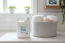 Load image into Gallery viewer, Walter & Rose laundry detergent bottle next to a basket of towels on a white surface with plants in the background.