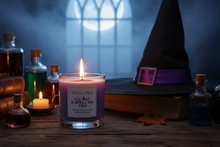 Load image into Gallery viewer, A lit purple candle in a glass jar sits on a wooden table surrounded by witch-themed props, including a black pointed hat with a purple band, old books, small bottles, and autumn leaves, with a glowing moonlit window in the background.