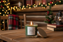 Load image into Gallery viewer, A lit green candle in a clear glass jar sits on a wooden table, with its silver lid leaning beside it. The background shows a cozy, blurred lodge interior with warm string lights, a small Christmas tree, and books.
