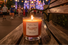 Load image into Gallery viewer, A lit candle in a clear glass jar sits on a wooden bench at night, with a Disney castle and walking guests blurred in the background. The label reads 'Walt Disney World Halloween Party' and 'The Magical Smoother Co.

