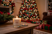 Load image into Gallery viewer, A lit scented candle in a clear glass jar sits on a wooden table in a cozy holiday living room, with a decorated Christmas tree, fireplace garland, and wrapped gifts blurred in the background.
